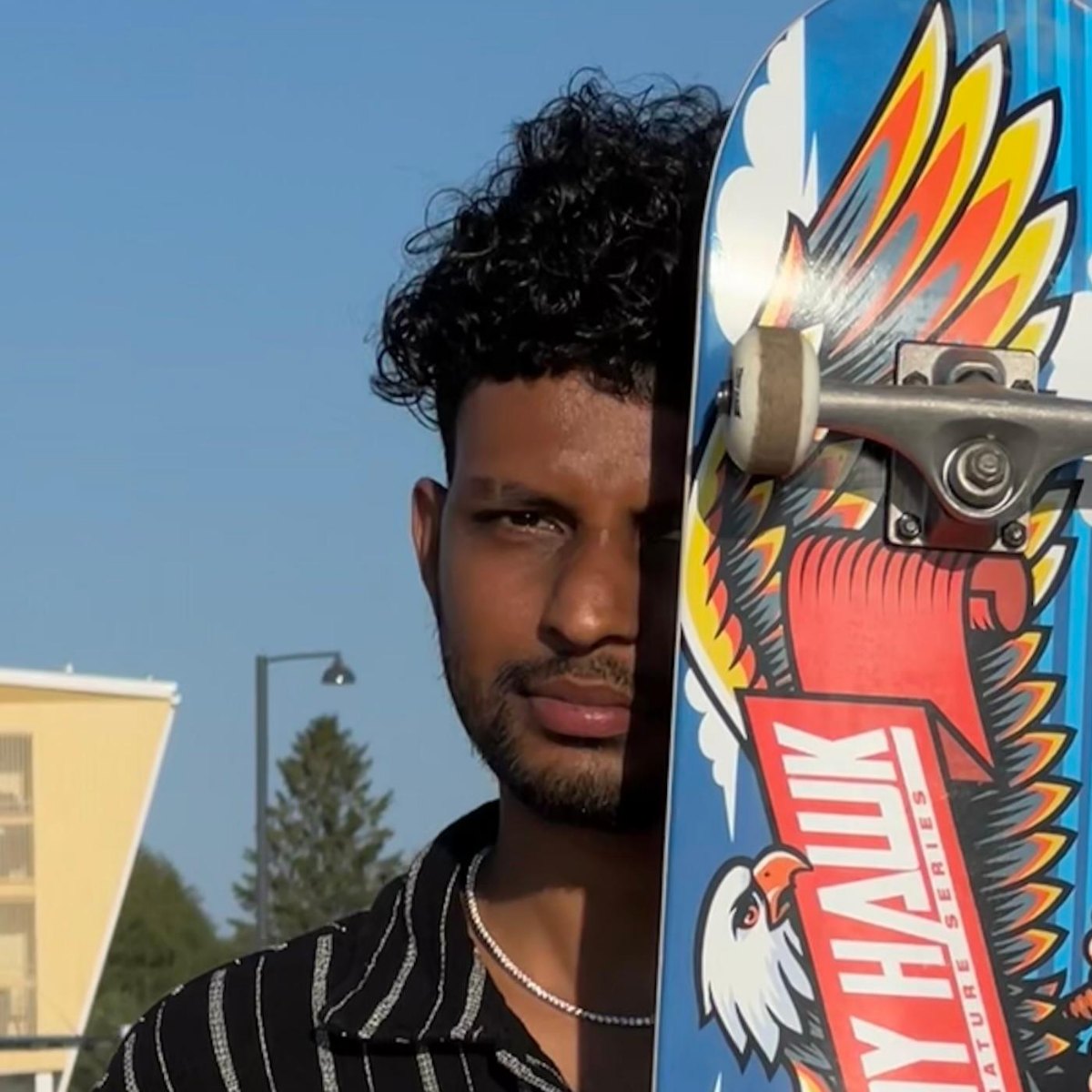 Kavindu Ravishan Perera outdoors in summer, holding a skateboard up over half his face