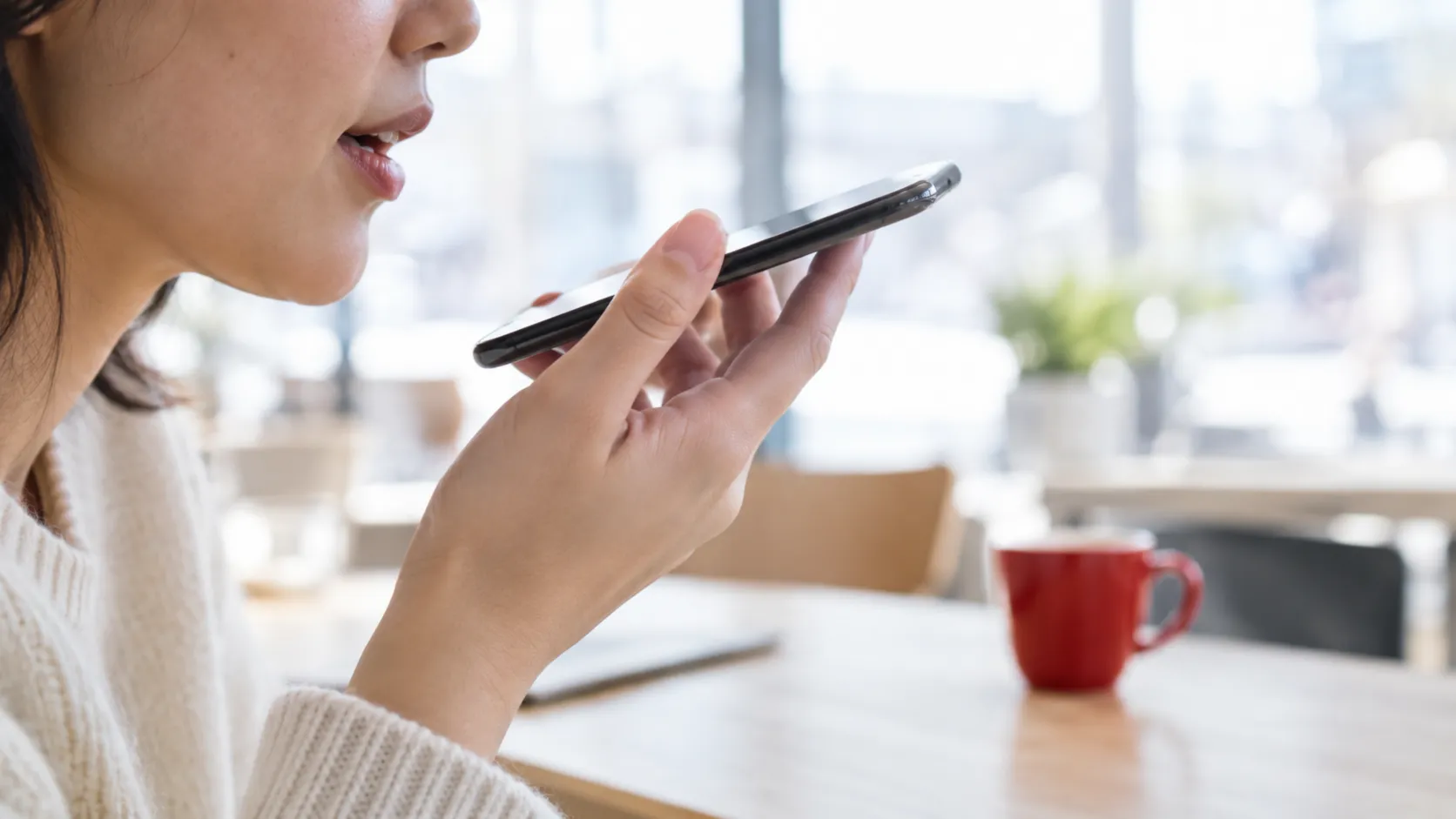 A person speaking into a phone in a bright cafe, a red cup on the table behind