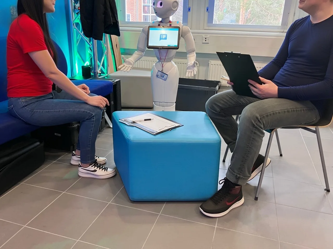 An interview in a small room: a participant and a researcher with a clipboard sit on either side of a low blue table, with Pepper standing between them.