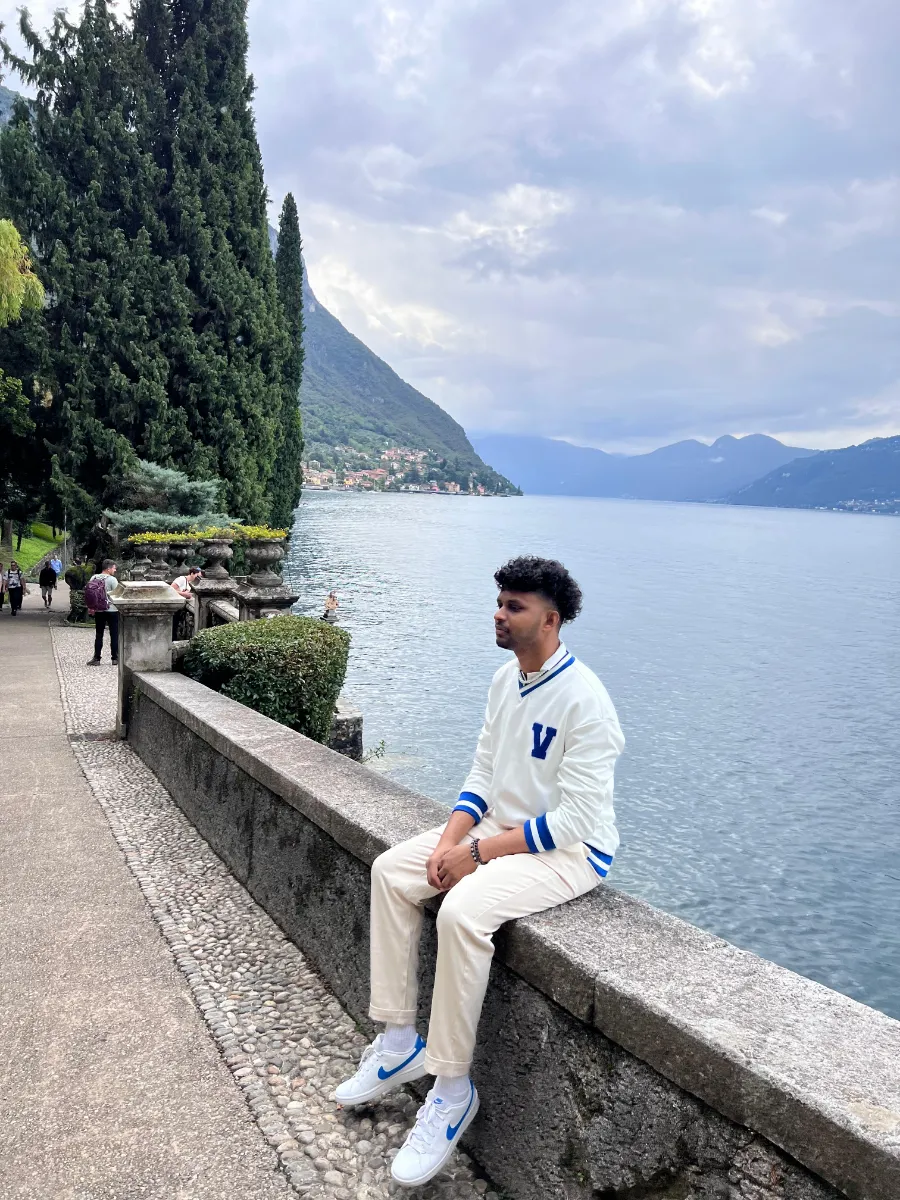 Kavindu Ravishan Perera sitting on a stone wall beside Lake Como, with mountains across the water